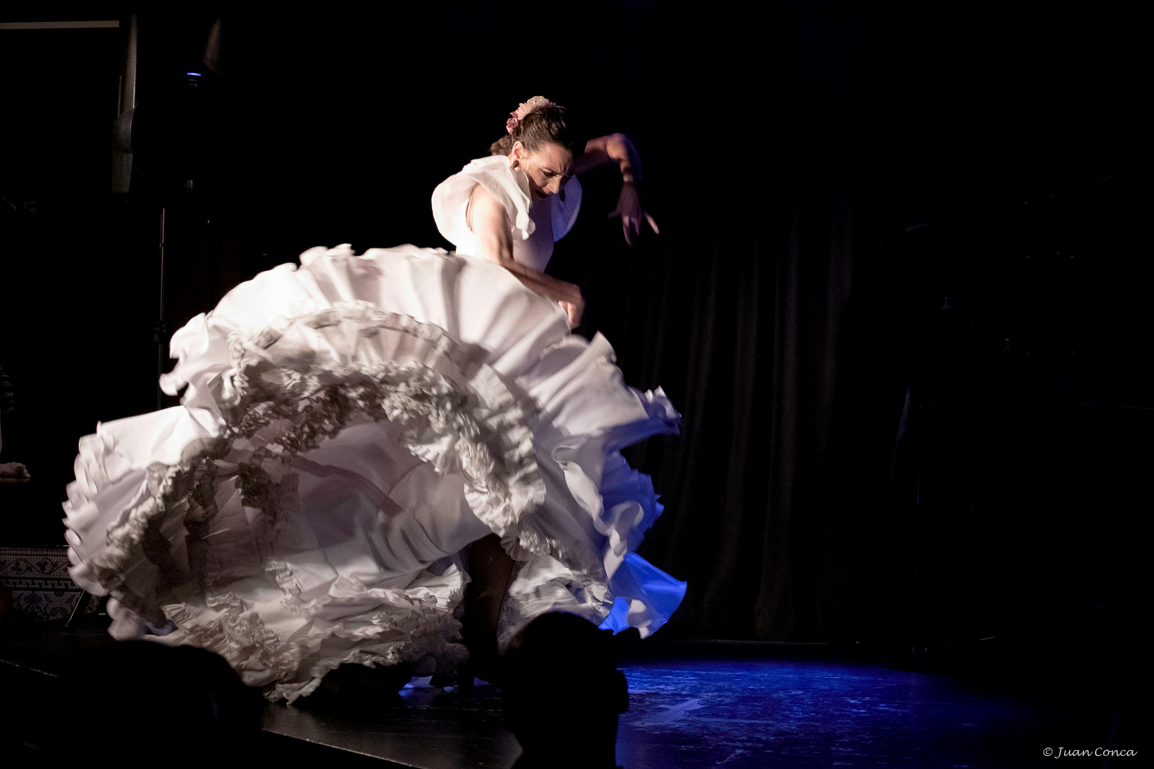 흰색 주름 장식 드레스를 입은 댄서가 어두운 커튼을 배경으로 어둡게 빛나는 무대에서 열정적으로 공연합니다.