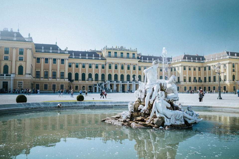 A grand yellow palace with many windows and a fountain featuring statues in the foreground, under a clear blue sky.