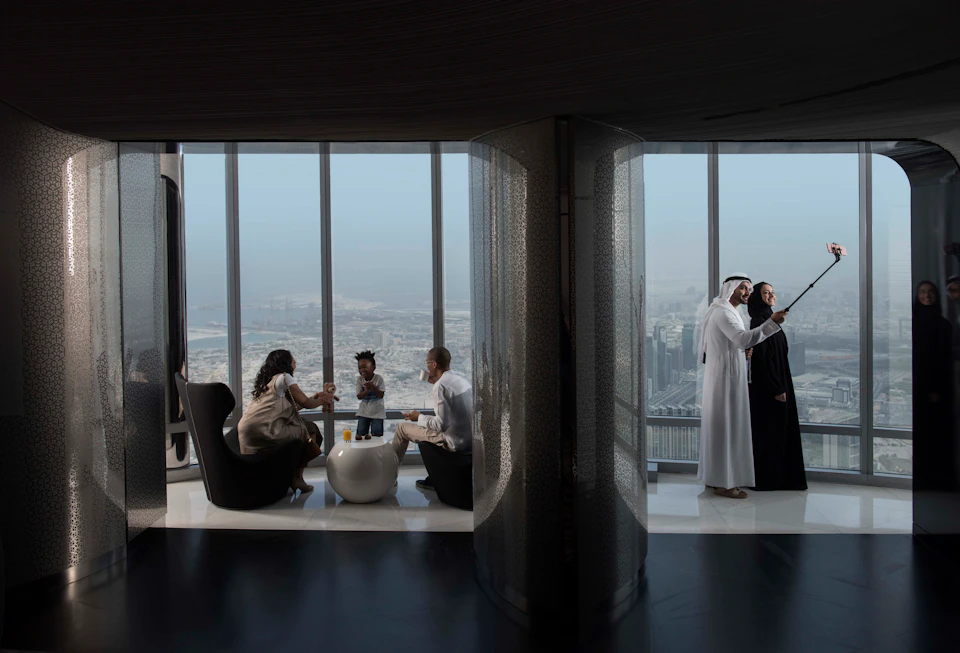 Two groups of people in a high-rise room with large windows showing a cityscape; one group seated, the other standing and talking.