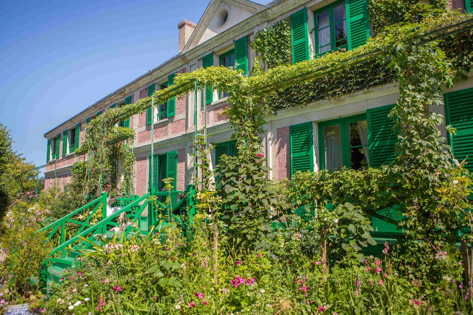 A building with green shutters and ivy-covered walls, surrounded by vibrant flowers and greenery on a sunny day.