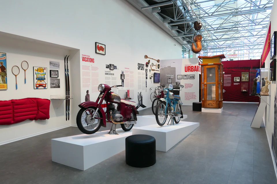 Museum exhibit featuring vintage motorcycles on white platforms, a yellow telephone booth, and wall displays with text and photos.