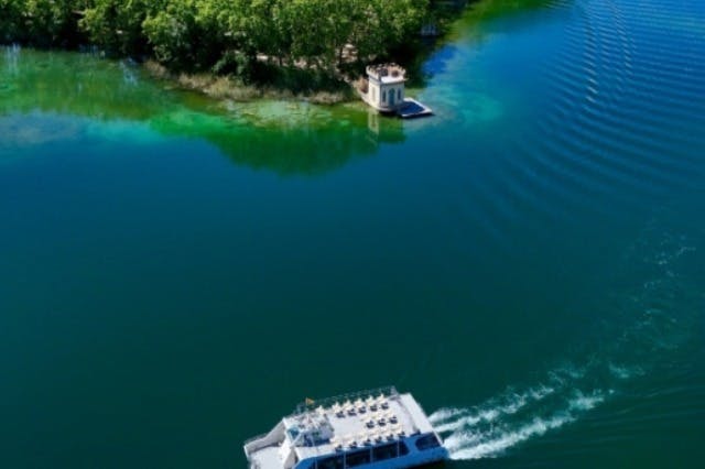 Vista aérea de um barco a deslocar-se num lago calmo perto de uma pequena ilha com árvores densas e uma pequena estrutura de pedra.