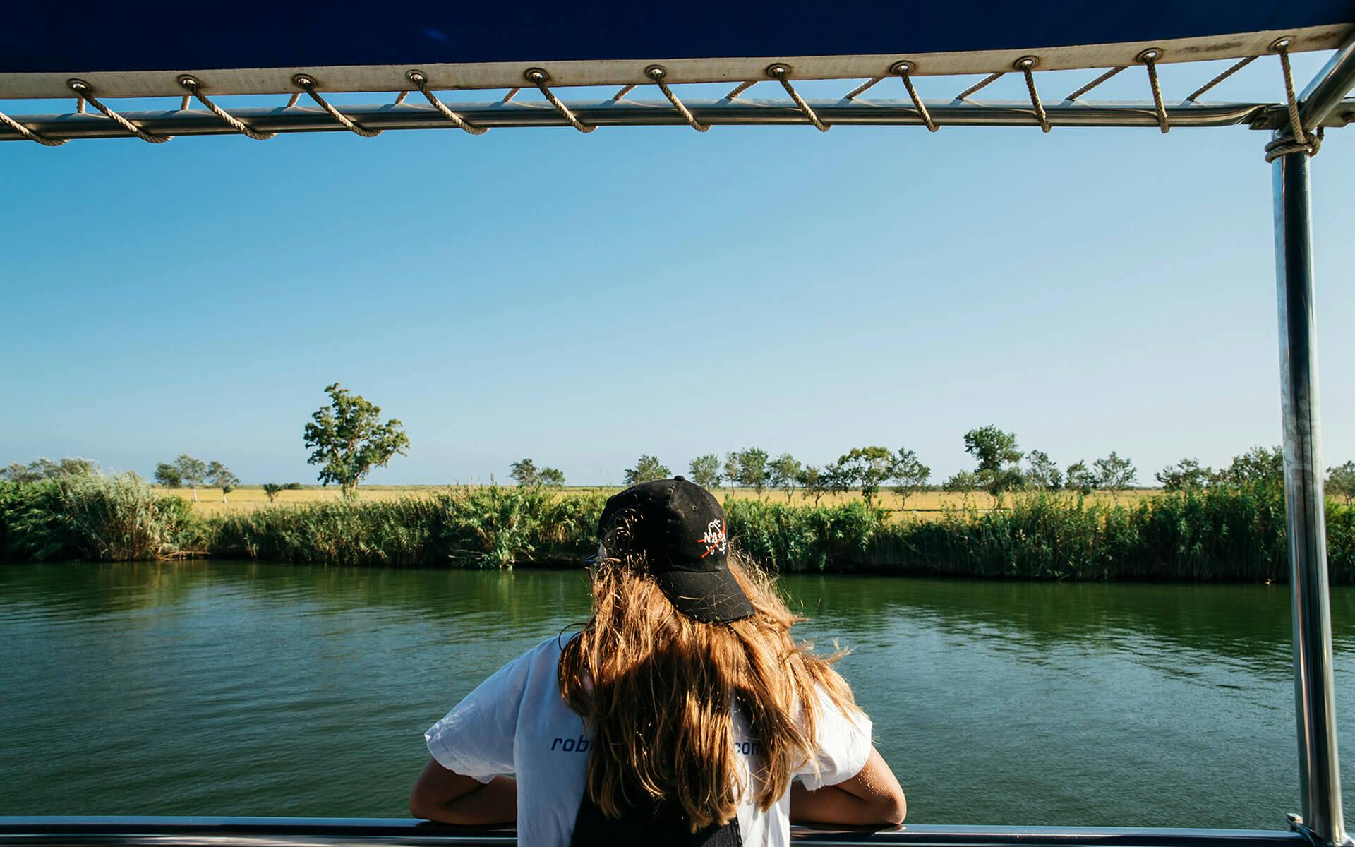 長い髪と黒い帽子をかぶった人がボートの上に立っていて、遠くに木がある穏やかな川と草むらの風景を眺めています。