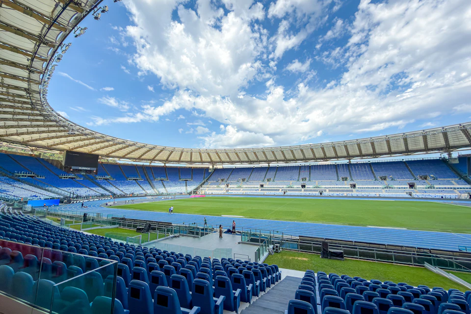 An empty stadium with blue seats, a green field, and a blue running track under a partly cloudy sky.