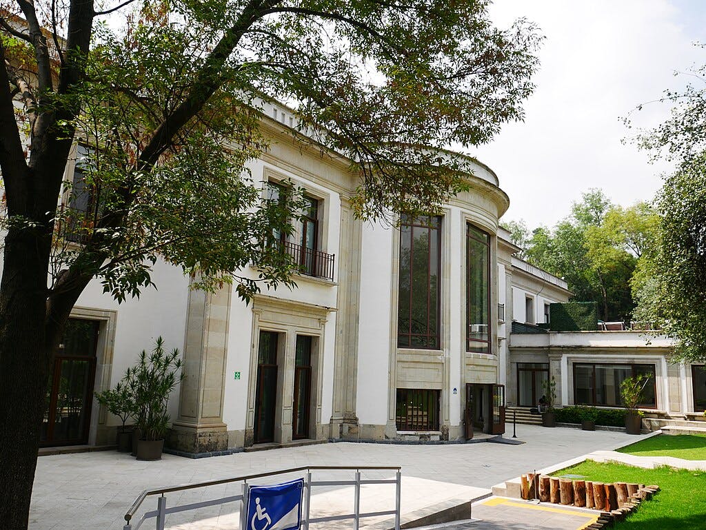 A white two-story building with large windows, surrounded by trees, a wheelchair ramp, and a courtyard with potted plants.
