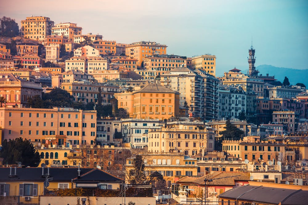 A densely packed hillside town with various colorful buildings under a slightly cloudy sky. A tall tower is visible in the distance.