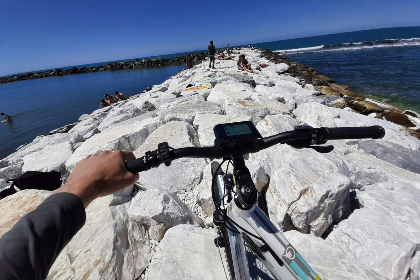 A person riding a bike on a rocky pier extending into the sea, with people sunbathing and the ocean in the background.