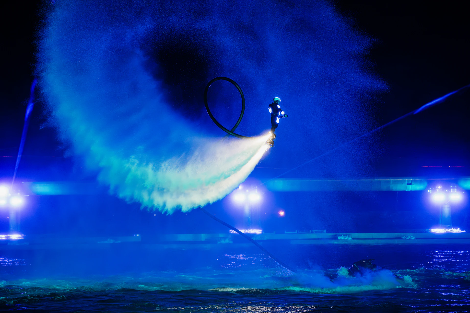 A person performing a nighttime water jetpack stunt, illuminated by blue lights, with a water spray forming a loop.