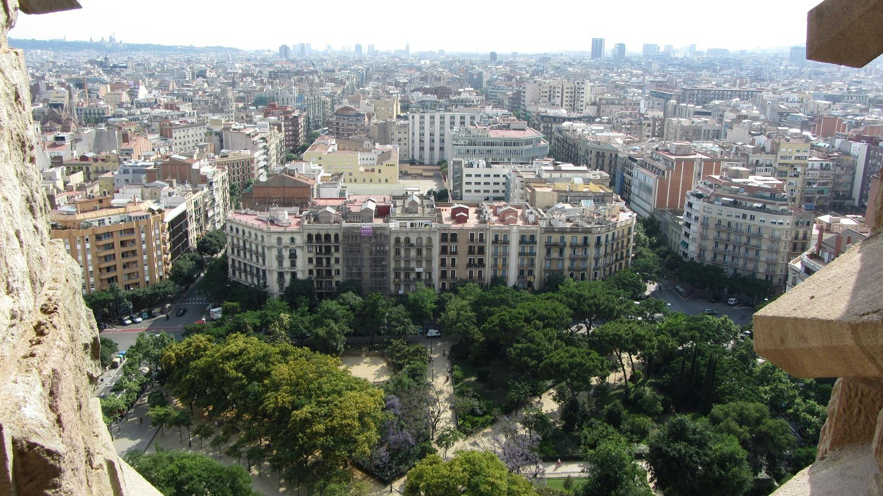 Aerial view of a densely built urban area with a large apartment building, surrounding greenery, and cityscape in the distance.