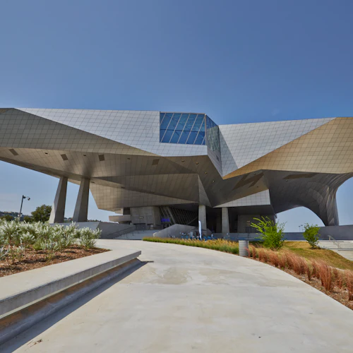 En modern byggnad med kantig, metallisk arkitektur, stödd av pelare, omgiven av anlagd grönska under en klarblå himmel.