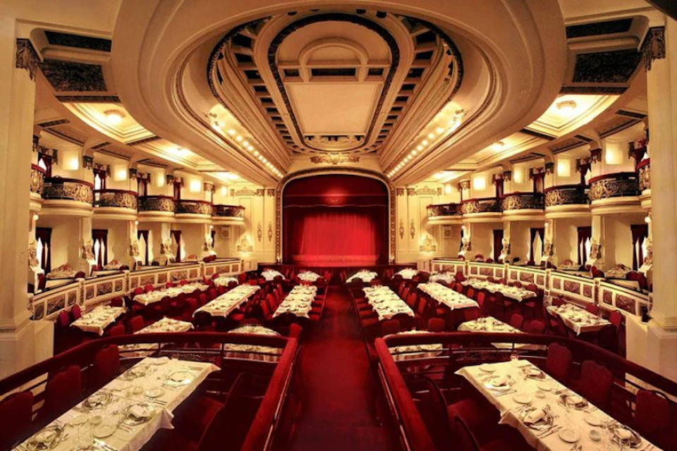 Elegant theater with a stage featuring a red curtain, surrounded by dining tables set with white tablecloths and place settings.