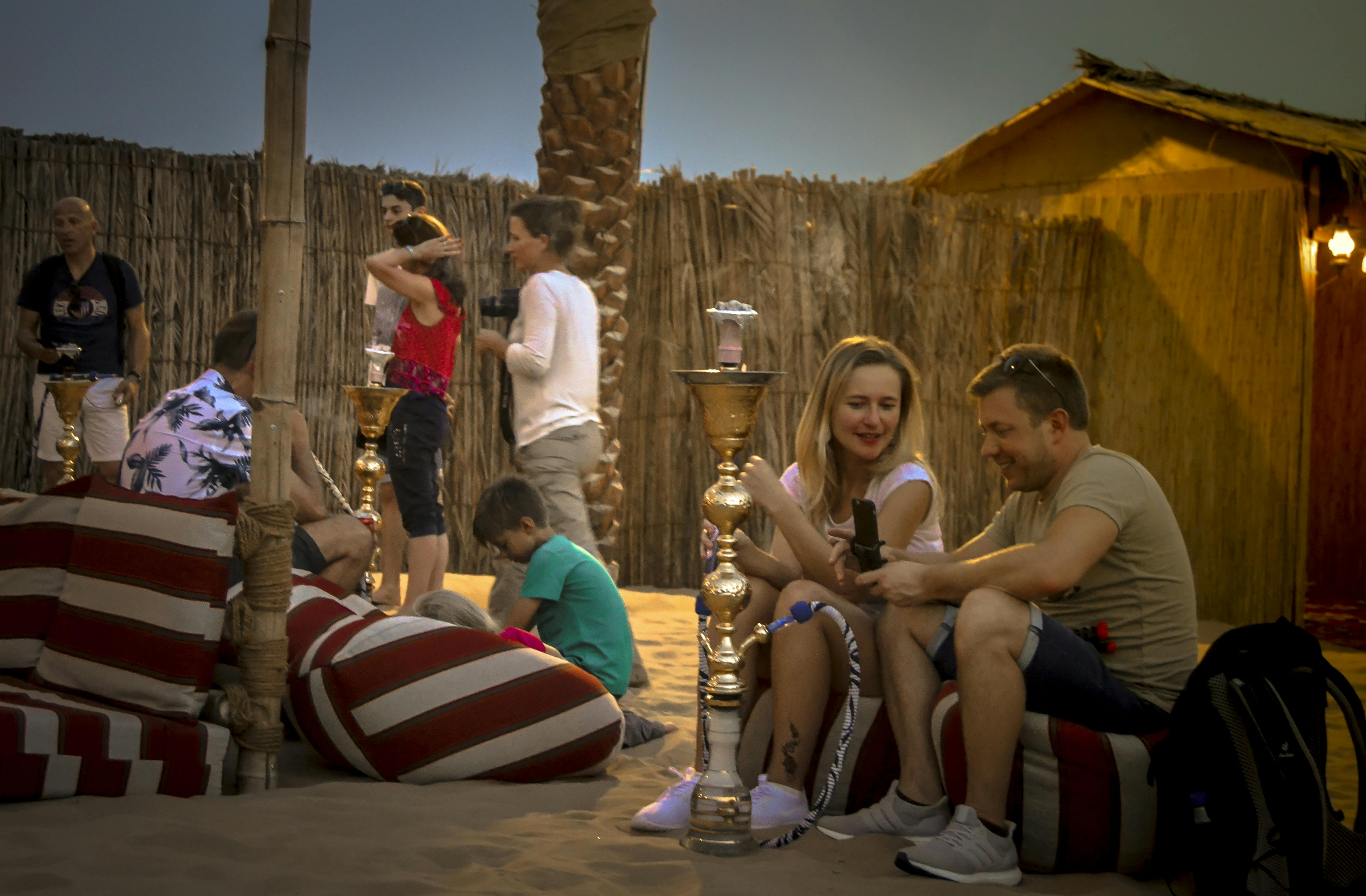 Lidé relaxují na pruhovaných polštářích v písčité oblasti s vodními dýmkami a dřevěným plotem v pozadí.