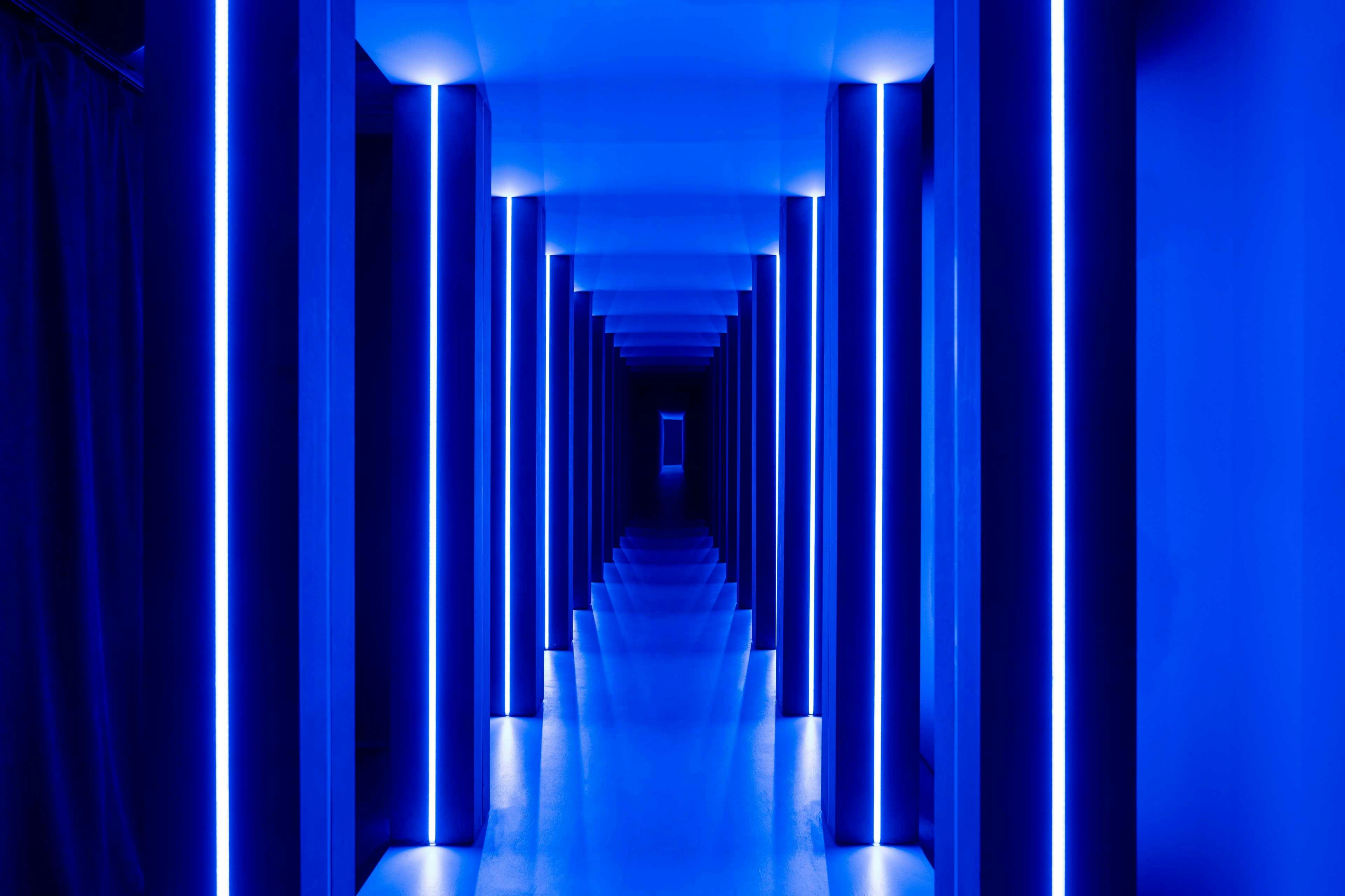 Ein langer, symmetrischer Flur mit vertikalen blauen Neonröhren an den Wänden, die eine futuristische und leuchtende Atmosphäre schaffen.