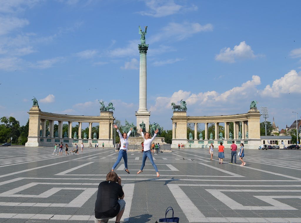 两个人跳到一个大广场上有雕像的纪念碑前，一个人蹲下来拍照。