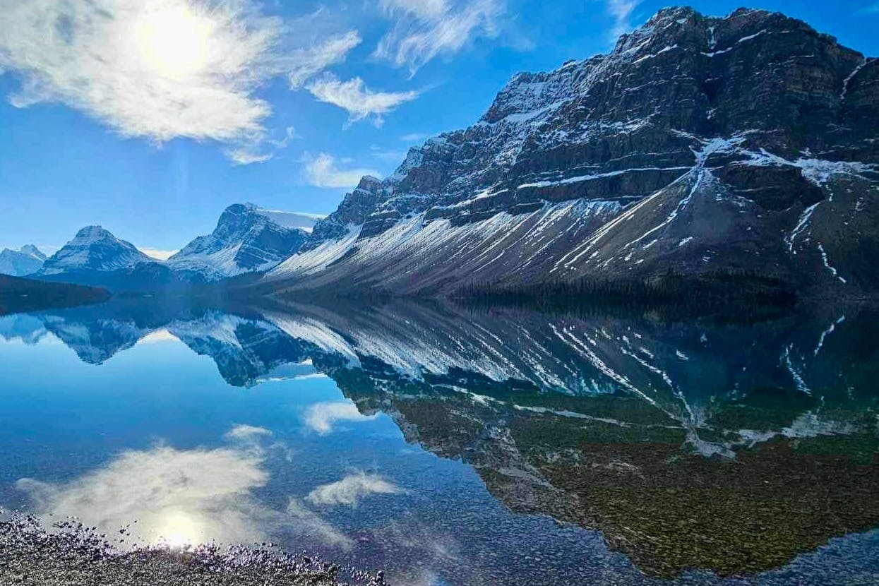 Snow-capped mountains reflected in a clear, still lake under a bright blue sky with scattered clouds.