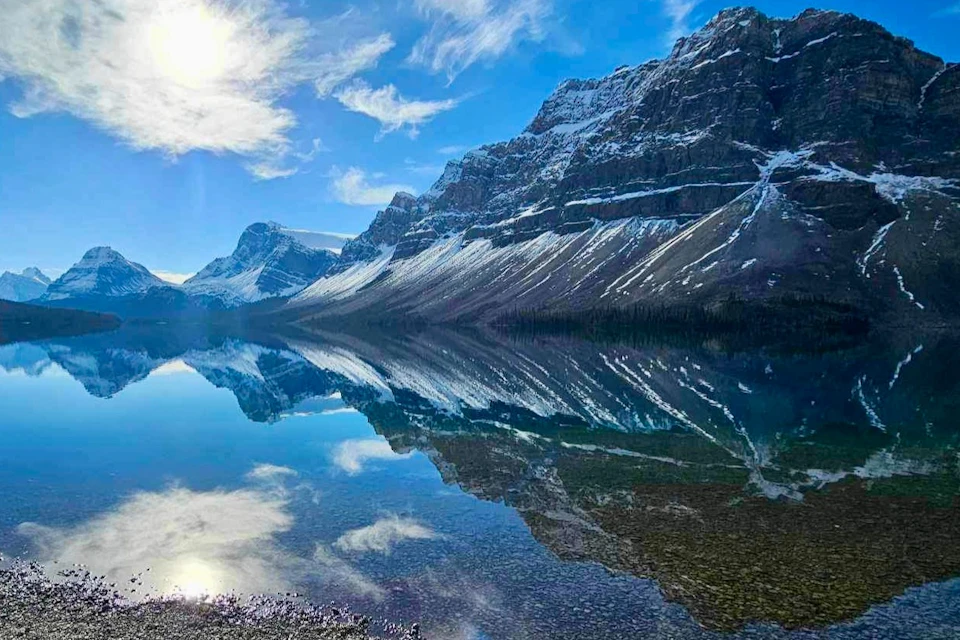 Snow-capped mountains reflected in a clear, still lake under a bright blue sky with scattered clouds.
