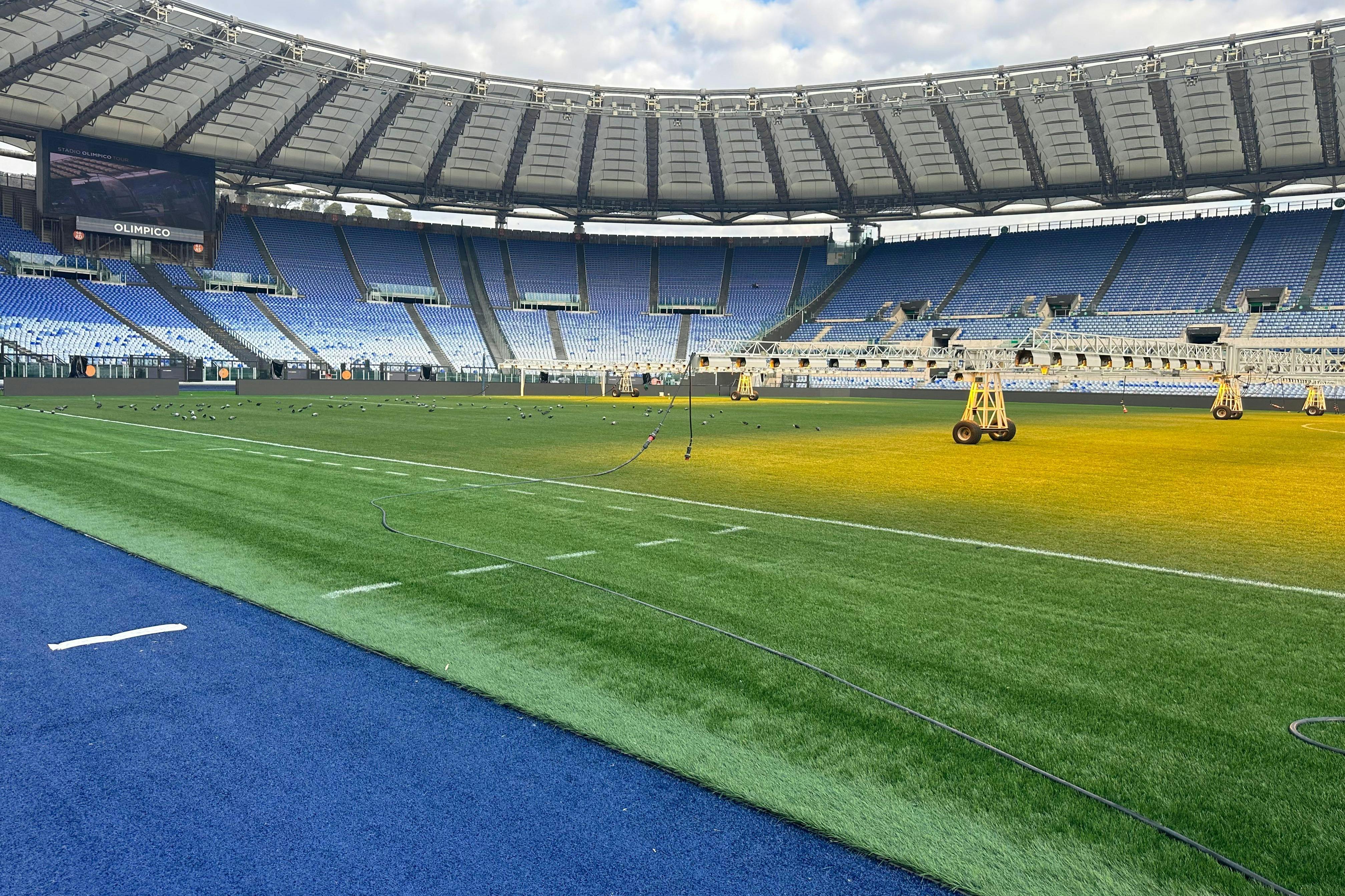 Ένα μεγάλο άδειο γήπεδο με μπλε καθίσματα και πράσινο πεδίο, με φορητό εξοπλισμό φωτισμού στο γρασίδι.