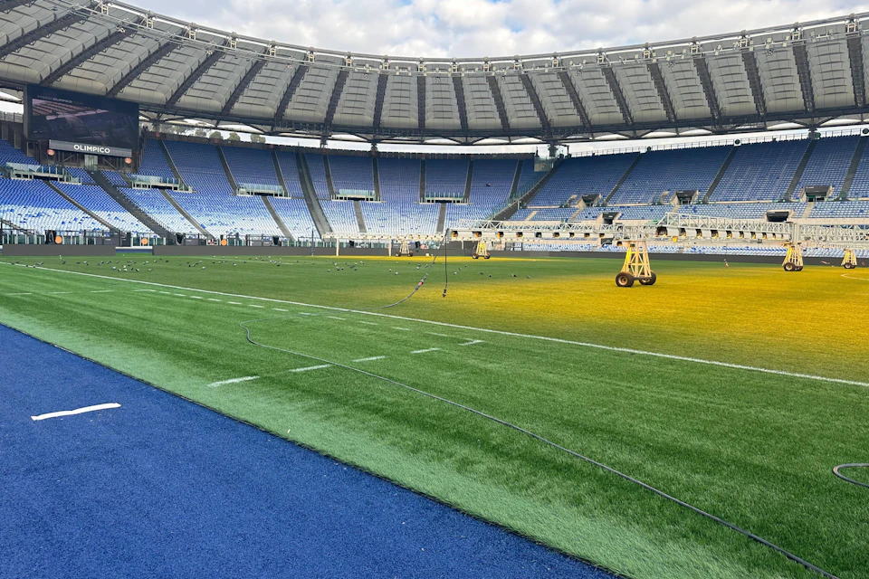 A large empty stadium with blue seats and a green field, featuring portable lighting equipment on the grass.