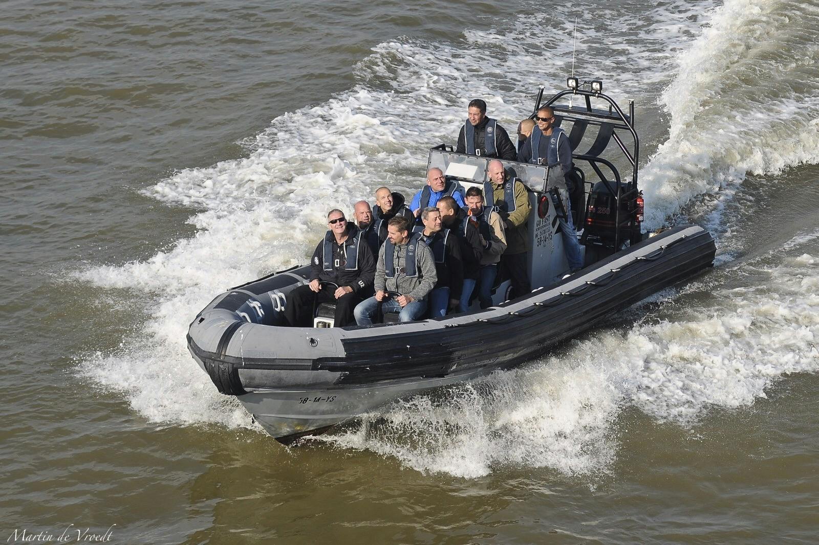 Devět lidí na motorovém nafukovacím člunu jede po rozbouřené vodě a vytváří za sebou bílou vlnovou stezku.