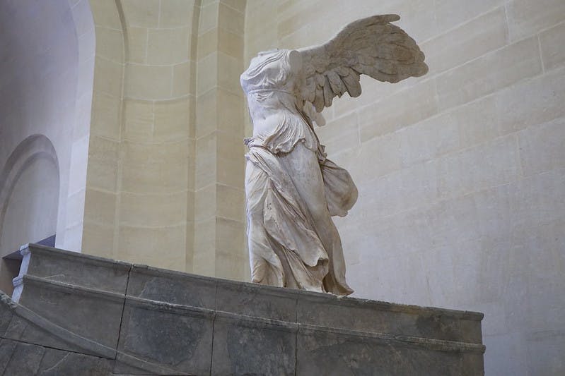 Το άγαλμα της Φτερωτής Νίκης της Σαμοθράκης εμφανίζεται σε μια πέτρινη πλατφόρμα σε μπεζ φόντο τοίχου.