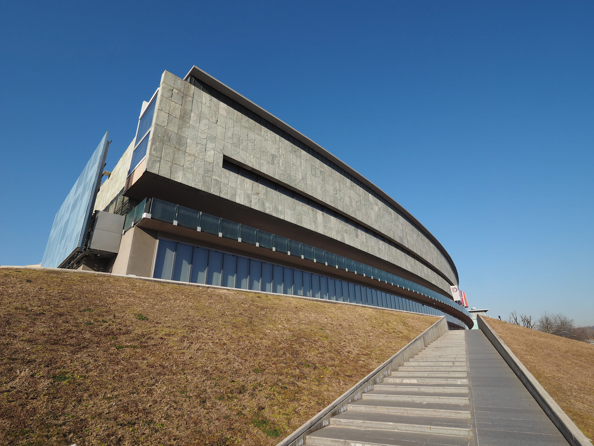 Modern, multi-story building with a curved facade and large windows, situated on a sloped grassy hill under a clear blue sky.