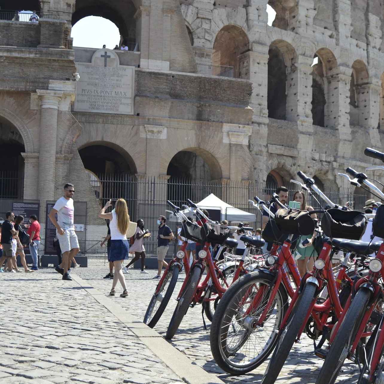 Tourists and rental red bikes in front of the Colosseum in Rome, with people taking photos and walking on a cobblestone path.