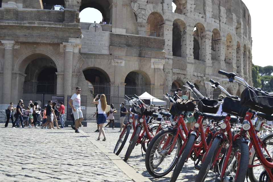 Tourists and rental red bikes in front of the Colosseum in Rome, with people taking photos and walking on a cobblestone path.