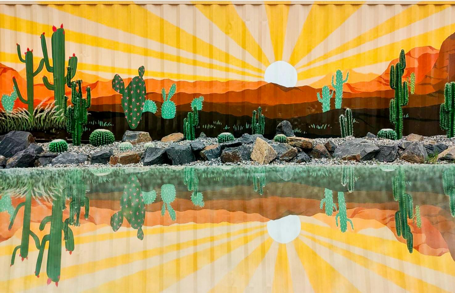 A mural of cacti in a desert sunset scene reflected in water, painted on a corrugated surface.