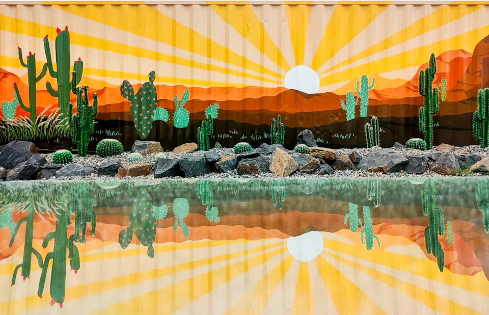 A mural of cacti and a sunrise with rocks and a reflective water surface in the foreground.