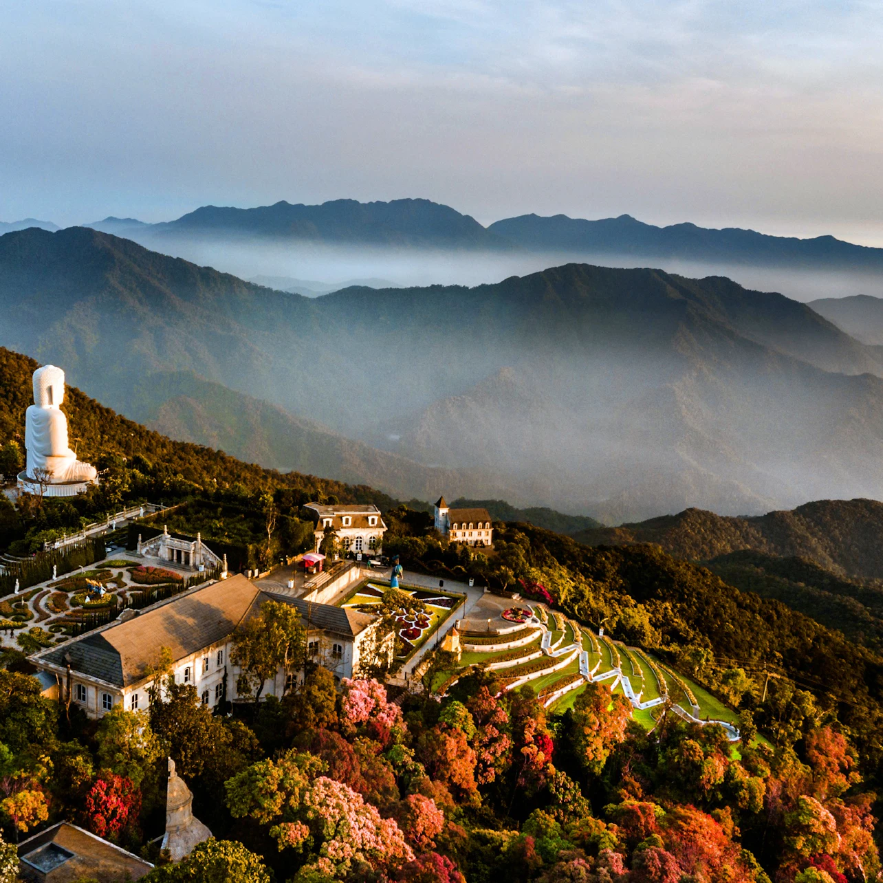 Aerial view of a mountain landscape with a temple complex, a large white statue, colorful trees, and misty hills in the background.