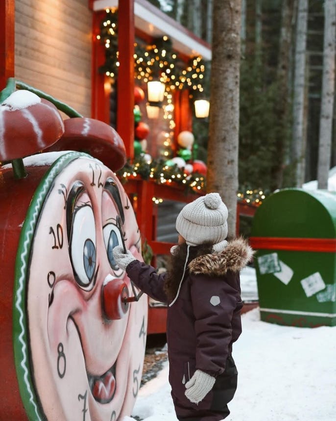 Een kind in winterkleding raakt een grote klok met een cartoonesk gezicht aan bij een versierd, feestelijk gebouw.