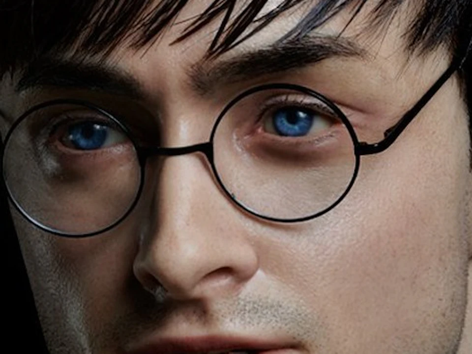 A person with short dark hair, wearing round glasses and a light-colored shirt, poses against a black background.