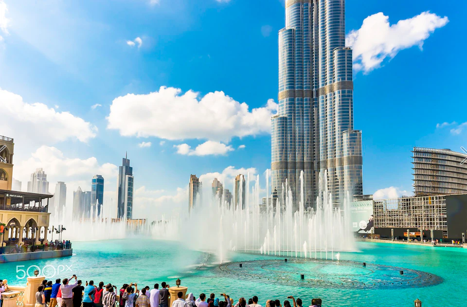 Tourists watch a large fountain show in front of a tall skyscraper on a clear, sunny day with a few clouds in the sky.