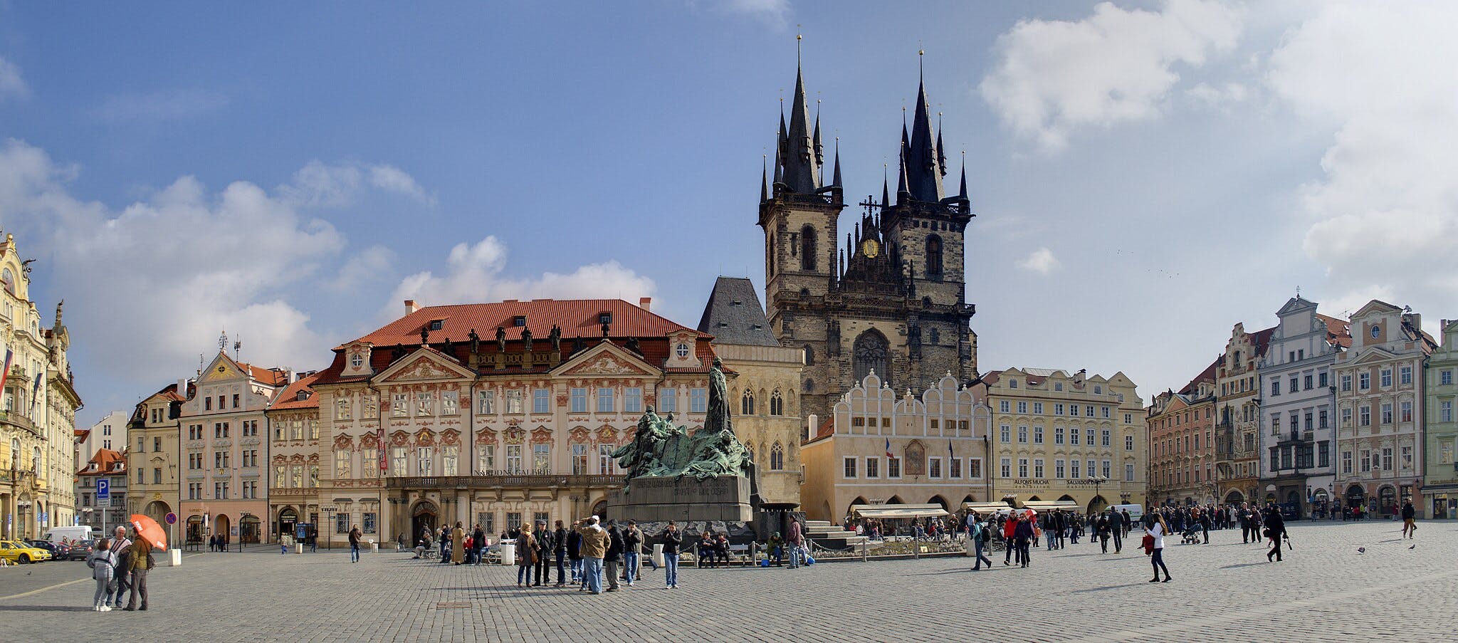 A bustling square with historical buildings, a large statue, and people walking and gathering under a sunny, partly cloudy sky.