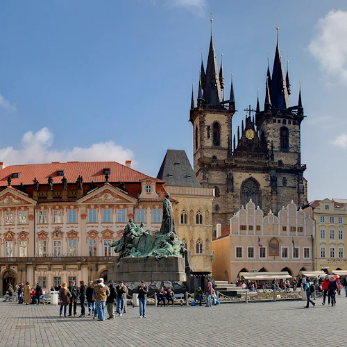 Ett livligt torg med historiska byggnader, en stor staty och människor som går och samlas under en solig, delvis molnig himmel.