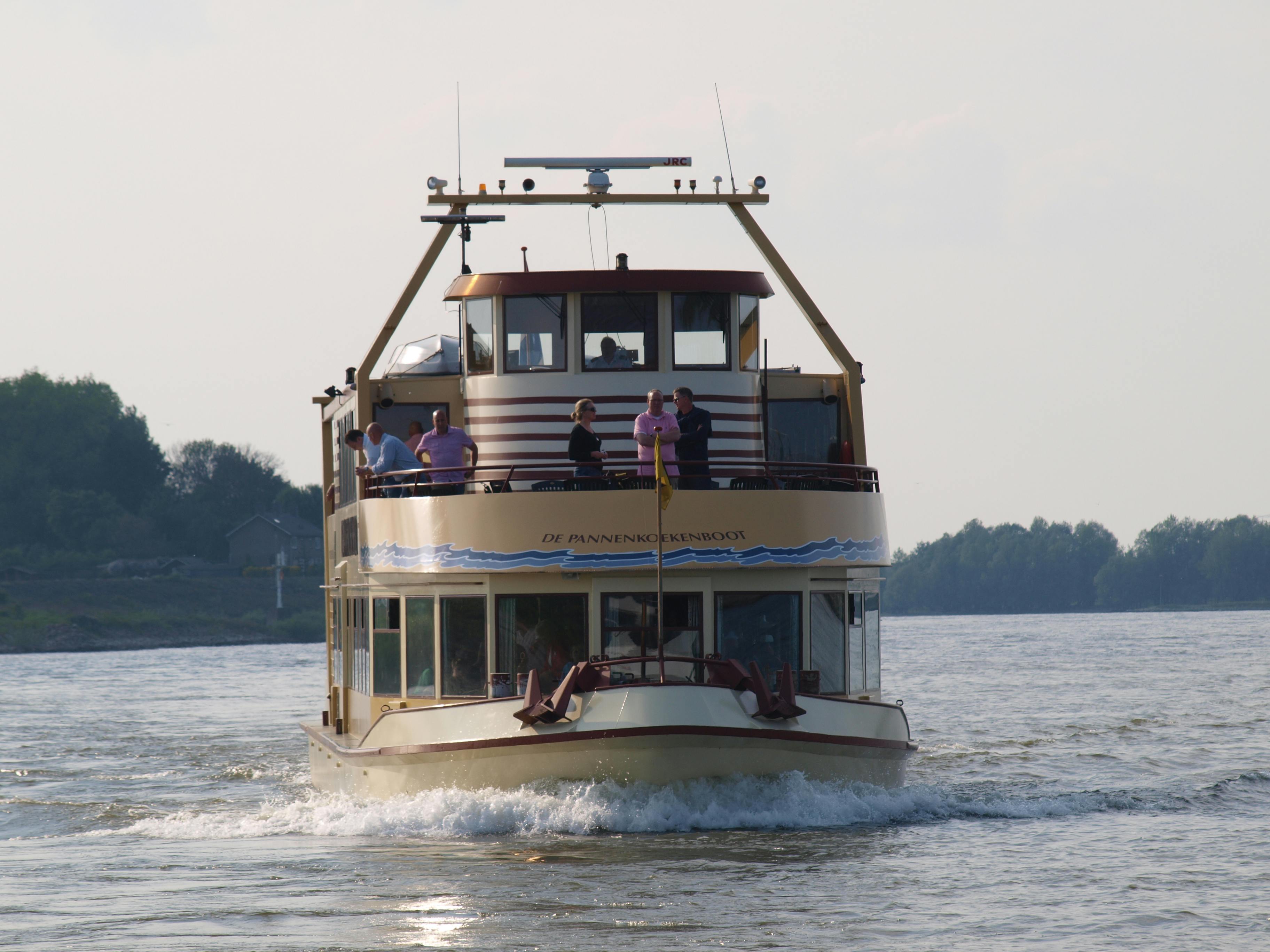 Barca a due livelli su un fiume con persone sul ponte superiore, alberi ed edifici sullo sfondo.