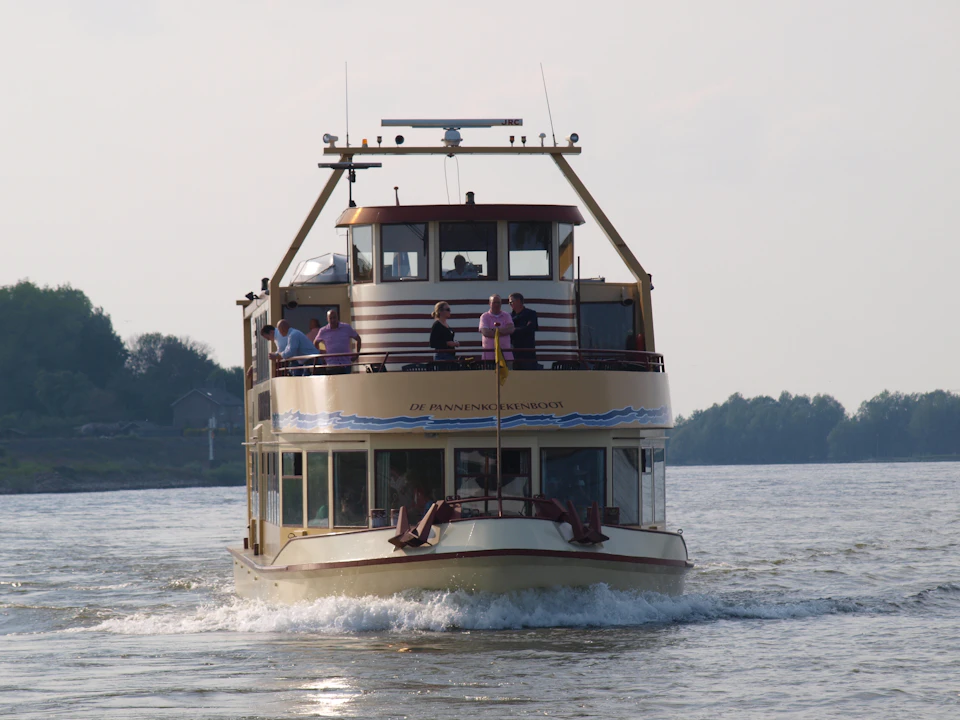 Two-tiered boat on a river with people on the upper deck, trees and buildings in the distant background.