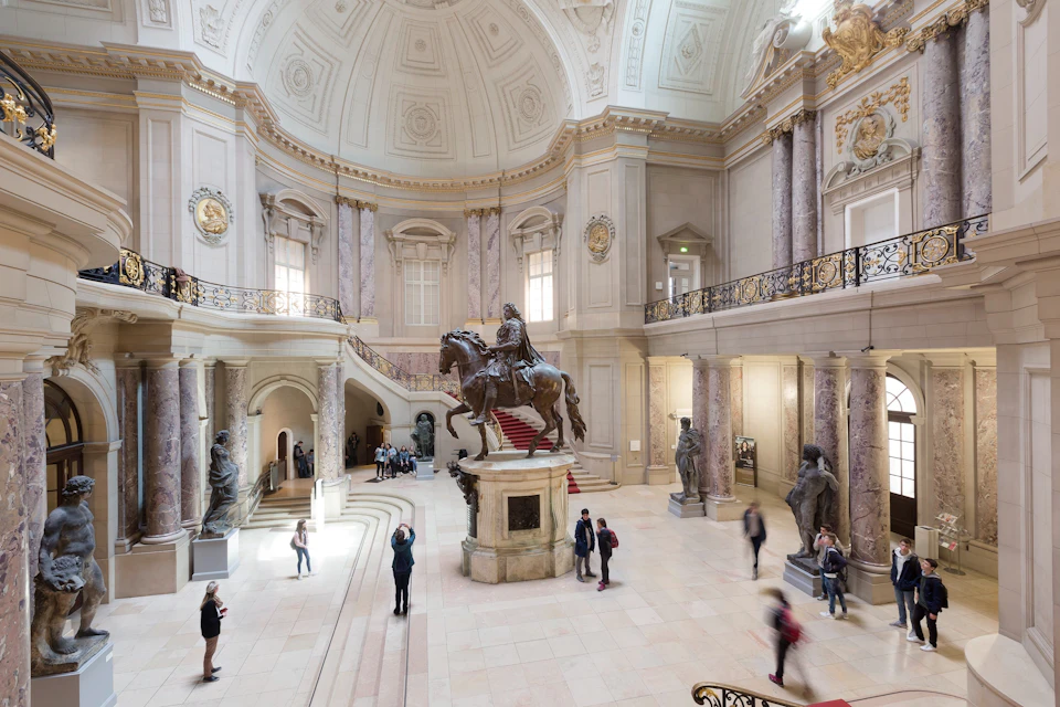 A grand hall with a statue of a rider on horseback, surrounded by visitors and ornate architectural details.