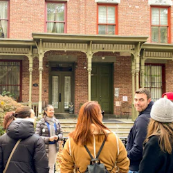 A group of people stand in front of a historic brick building with green doors and large windows, chatting and smiling.