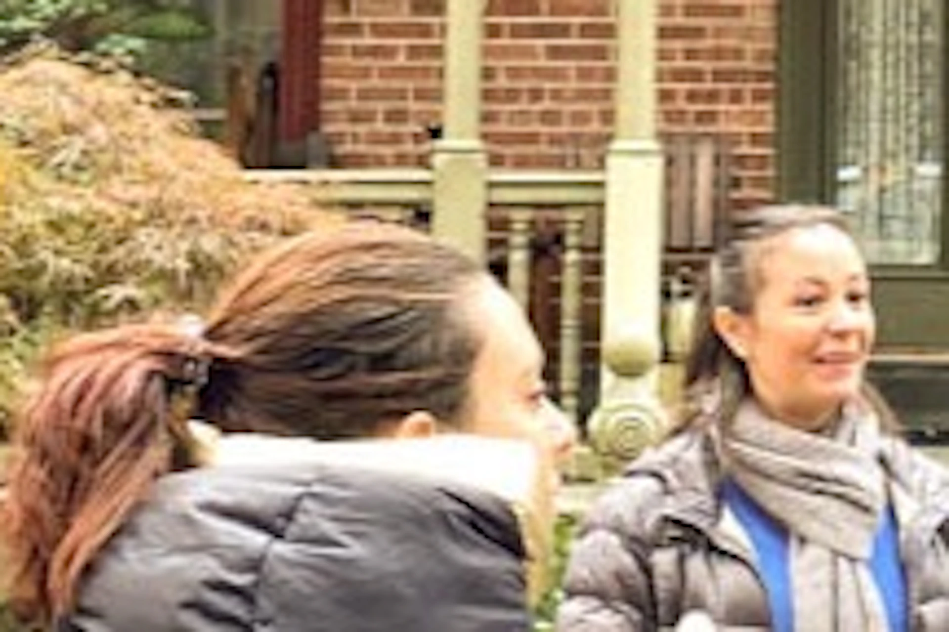 A group of people stand in front of a historic brick building with green doors and large windows, chatting and smiling.