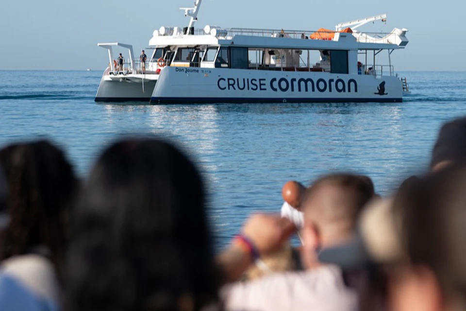 A catamaran labeled 'CRUISE CORMORAN' on calm water, with blurred figures in the foreground.