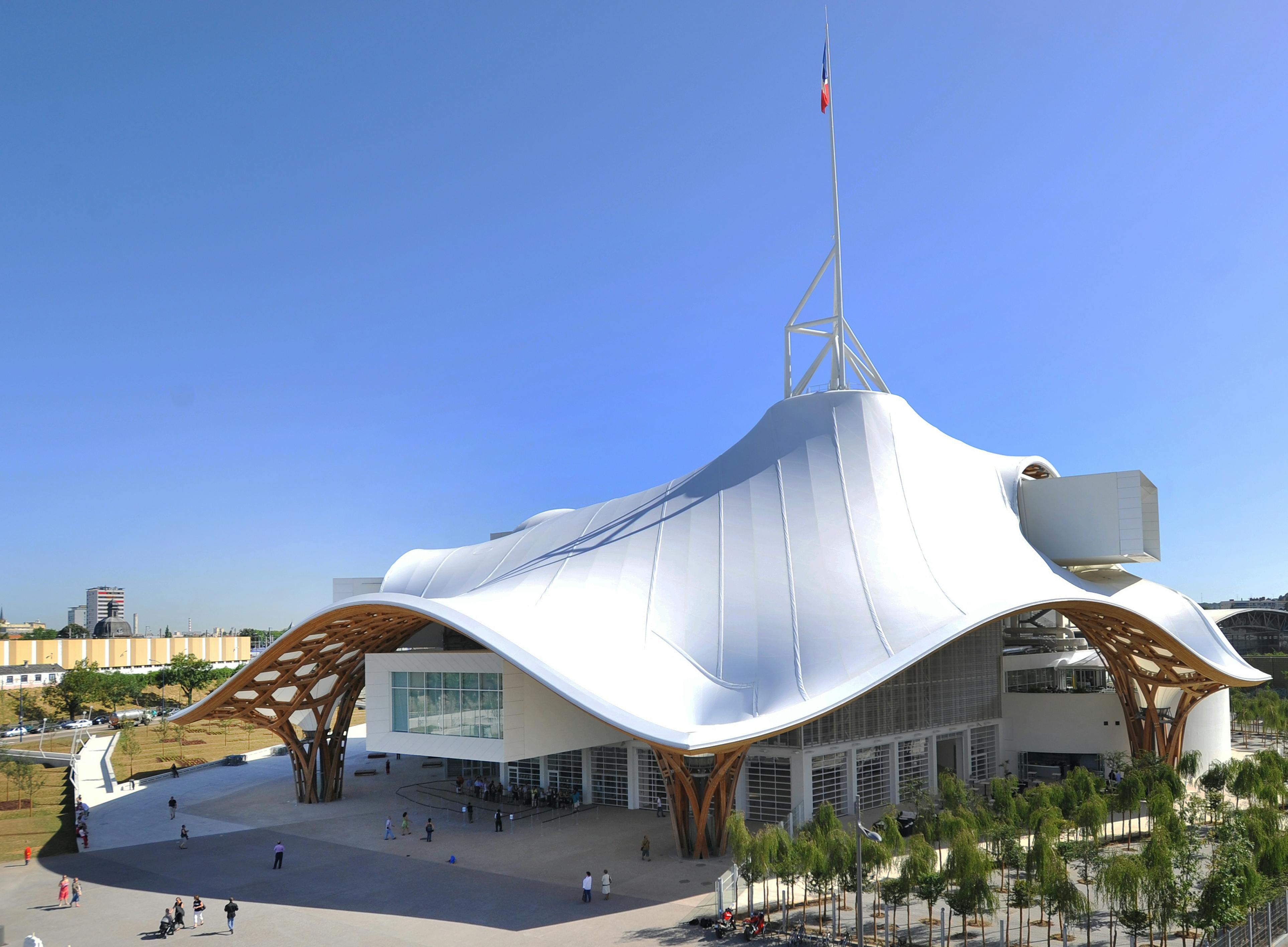 Modern building with an undulating white roof, wooden supports, and a tall flagpole, surrounded by people and greenery under clear blue sky.