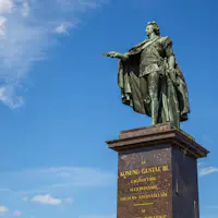 Bronzen beeld van een man in antieke kleding op een hoge sokkel met gegraveerde tekst tegen een blauwe lucht met pluizige wolken.