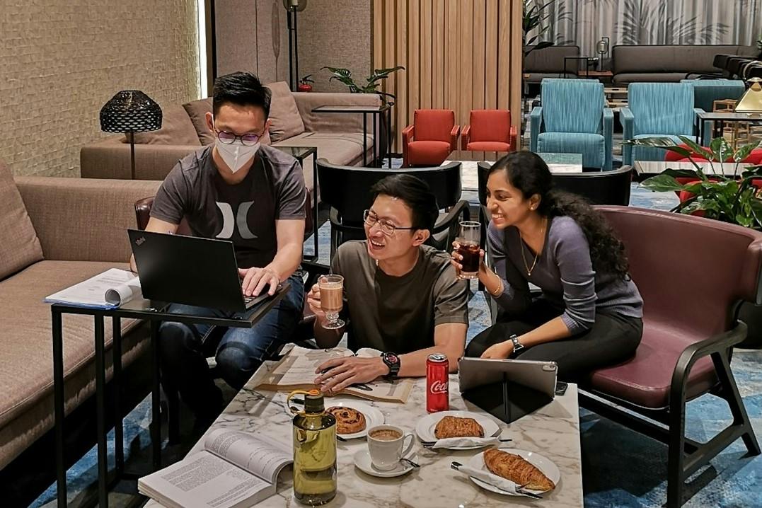 Three people gathered at a table with laptops, books, and beverages, smiling and engaging in a casual discussion.