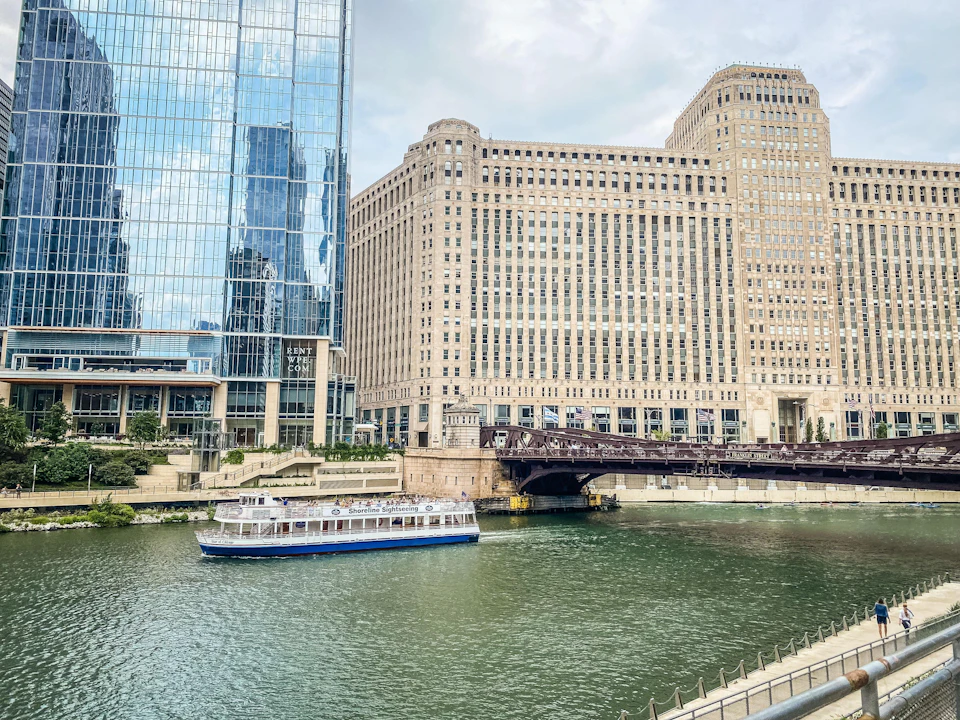 A tour boat navigates a river near a large glass building and a historic stone building with a bridge in the background.