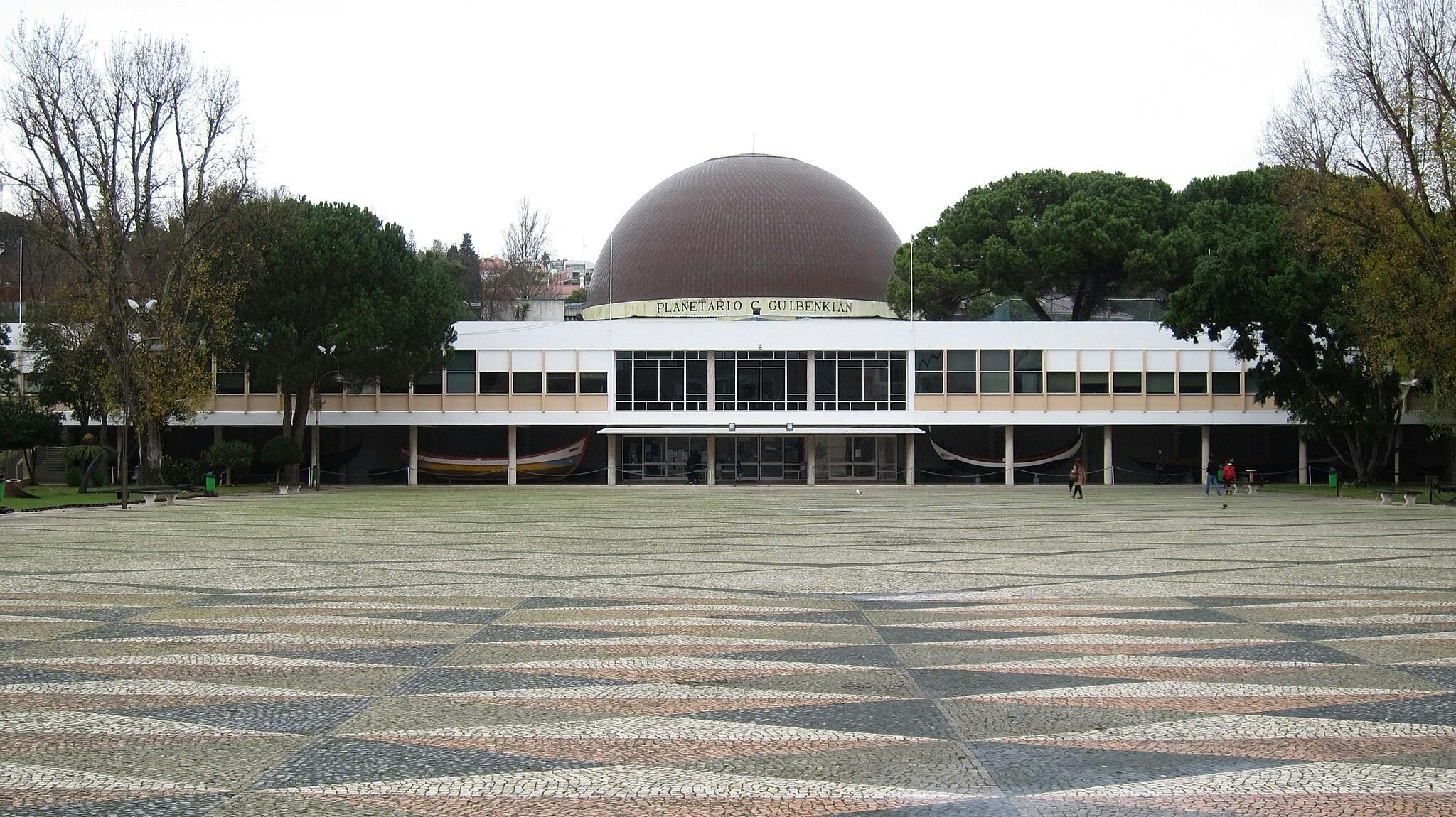 Large, dome-shaped planetarium building with "Planetário C Gulbenkian" sign, surrounded by trees and geometric-patterned plaza. Few people walking.