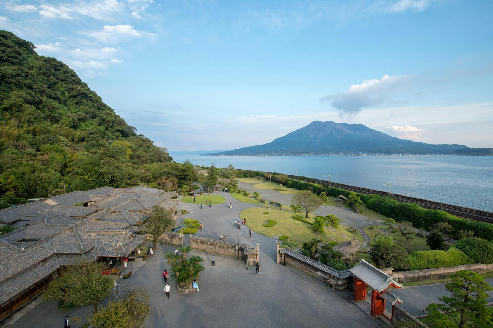 Scenic view of a park with trees, a traditional building, and a mountain across the water under a clear blue sky.