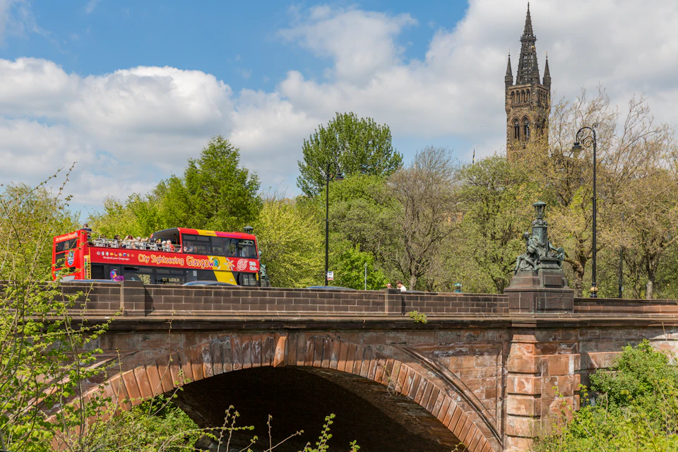 Red double-decker tour bus crossing a stone bridge with a tall church spire and trees in the background on a sunny day.