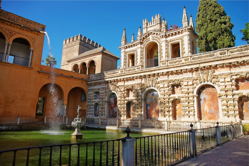 A historic building with ornate architecture and a fountain in a green pond, surrounded by a railing under a clear blue sky.