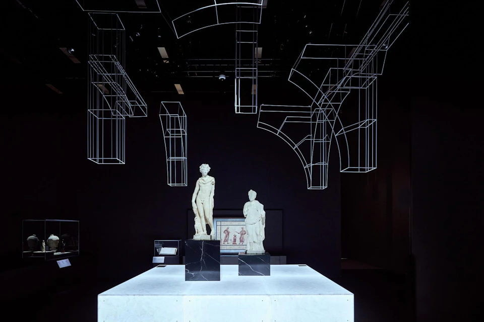 Two classical statues on a lit pedestal in a dark room, with wireframe structures suspended above.